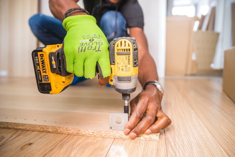 A worker in a work glove driving a screw into a floor bracket with a cordless impact driver.